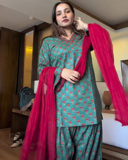 A woman modeling a teal green and pink printed salwar kameez set with a bright pink dupatta in a hotel room.