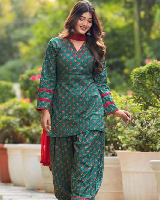 A woman wearing a green and red floral print salwar kameez suit with a red dupatta, standing in a garden setting.