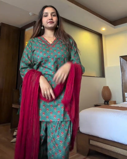 A woman modeling a green and pink patterned kurta set with a bright pink dupatta in a hotel room setting.