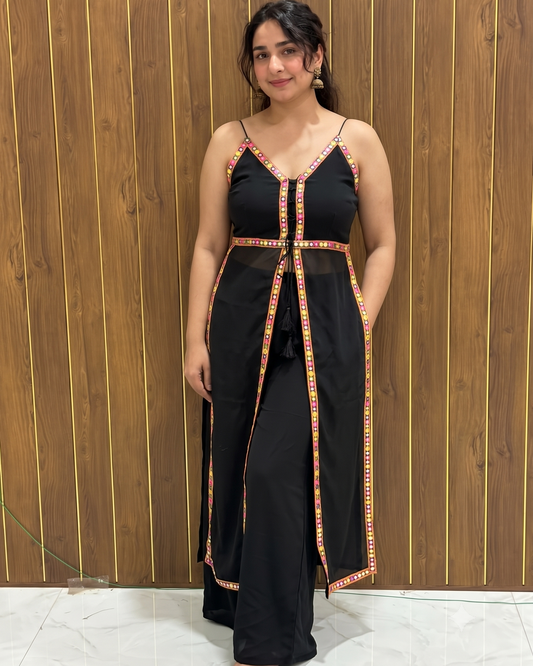 A woman modeling a black maxi dress with red and yellow patterned trim along the straps, neckline, waist, and front drape, standing against a brown wood-paneled wall.