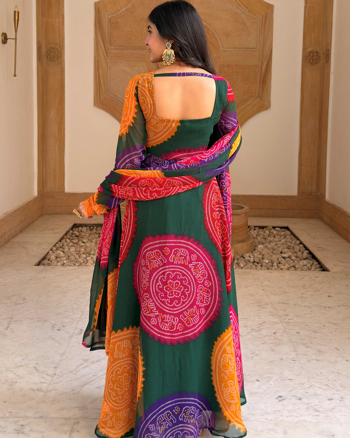 A woman stands with her back to the camera wearing a dark green saree with vibrant pink, orange, and purple circular mandala patterns, set against a traditional indoor background with wooden wall panels.
