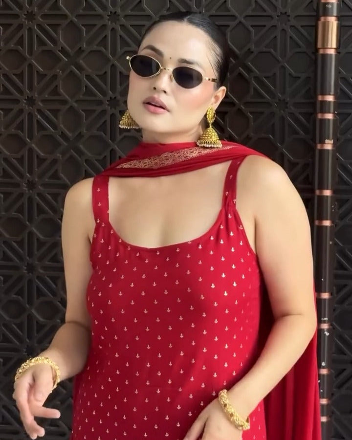 Woman in a red dress with a pattern, sunglasses, and gold jewelry against a dark textured wall.
