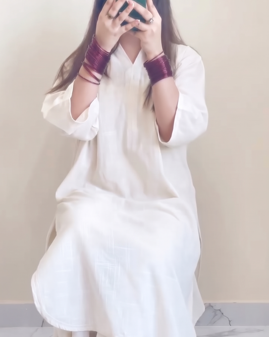 A person wearing a loose white tunic dress with matching white pants and dark brown sandals, seated against a plain beige wall with a marble floor.